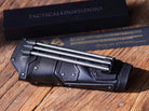 A black leather Kirin Vambrace with slots containing three Bo-Shurikens, displayed on a wooden surface next to its packaging.